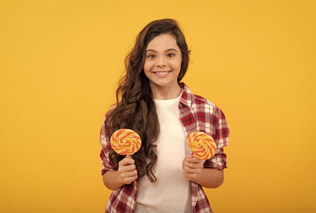 Niño feliz con el pelo largo y rizado en camisa sostenga caramelo de paleta sobre fondo amarillo delicioso