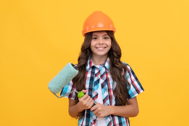 Niño feliz con el pelo rizado en el casco de la construcción mantenga la pintura del rodillo de pintura