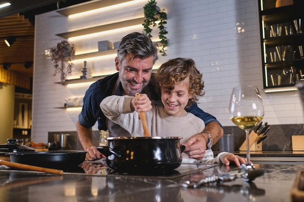 Foto niño feliz de pelo rizado revolviendo un plato caliente en una cacerola y ayudando a un padre de mediana edad a cocinar mientras pasan tiempo juntos en la cocina de casa