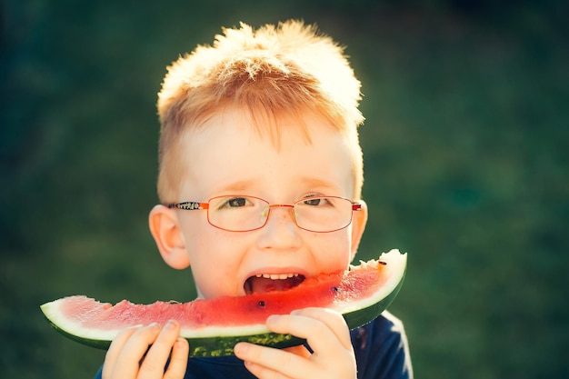 Niño feliz con pelo rojo en camisa azul y anteojos en cara sonriente feliz comiendo sandía día de verano al aire libre sobre fondo verde natural