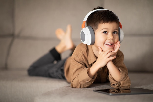 Un niño feliz y pensativo con auriculares inalámbricos piensa que una idea creativa viendo un video en una tableta se encuentra en el sofá