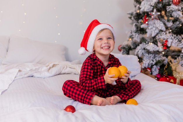 niño feliz en pijama y gorro de Papá Noel en casa en la cama comiendo mandarinas para Navidad