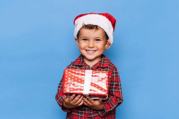 Un niño feliz en pijama de Navidad y un sombrero con una caja de regalo. la foto de estudio está dorada sobre un fondo azul. El concepto de vacaciones.