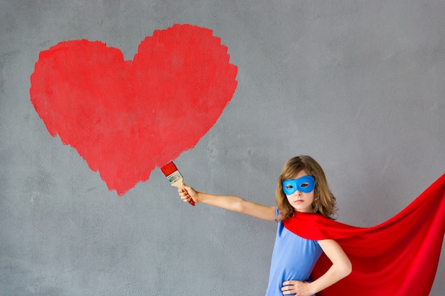 Niño feliz pintando gran corazón rojo en la pared. Chica divertida superhéroe jugando en casa. Tarjeta del día de San Valentín. Concepto de renovación y diseño