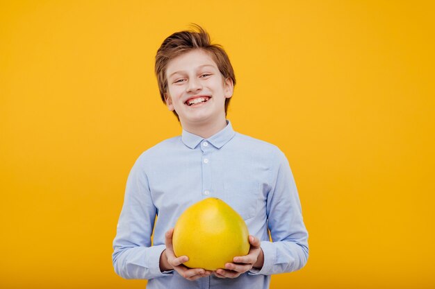 Niño feliz preadolescente con fruta de pomelo en la mano, con la camisa azul, aislado en la pared amarilla