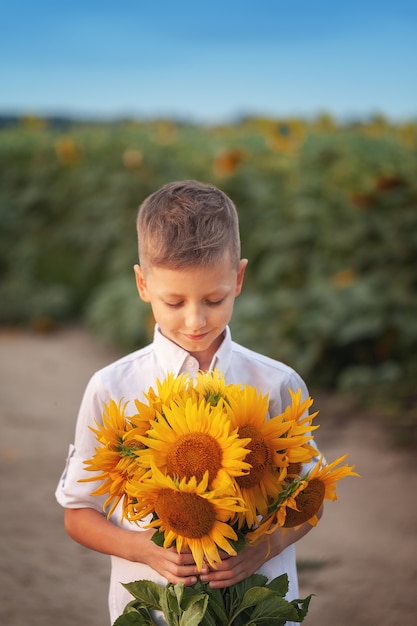 Niño feliz con el ramo de girasoles hermosos en campo del girasol del verano en puesta del sol. Día de la Madre