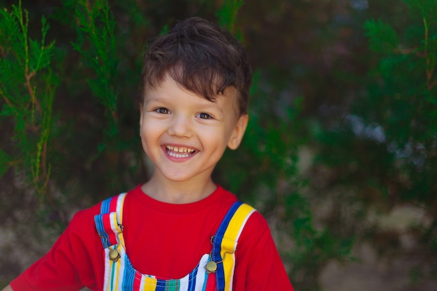 El niño feliz se ríe. Un niño con una camiseta roja y un mono brillante en la calle, sobre un fondo verde de abeto.