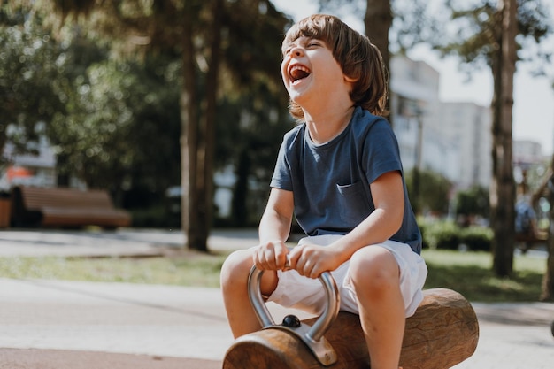 Un niño feliz y riéndose con una camiseta azul y pantalones cortos blancos monta en una mecedora de madera al aire libre. el niño está jugando en un parque público en un día soleado de verano. estilo de vida. espacio para texto