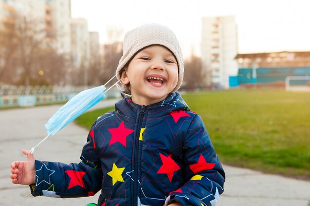 Niño feliz y risueño en un paseo por un parque de la ciudad le quita una máscara médica de la cara.