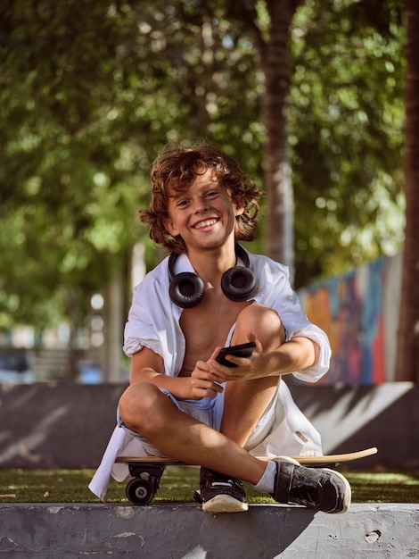 Foto niño feliz con ropa informal y auriculares sonriendo y mirando a la cámara mientras está sentado en patineta con las piernas cruzadas en un banco en el parque y con un teléfono celular en las manos en un día soleado