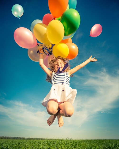 Niño feliz saltando con coloridos globos de juguete al aire libre. Niño sonriente divirtiéndose en el campo de primavera verde contra el fondo del cielo azul. concepto de libertad