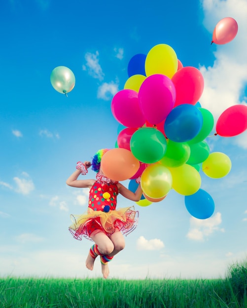 Niño feliz saltando con coloridos globos de juguete al aire libre. Niño sonriente divirtiéndose en el campo de primavera verde contra el fondo del cielo azul. concepto de libertad