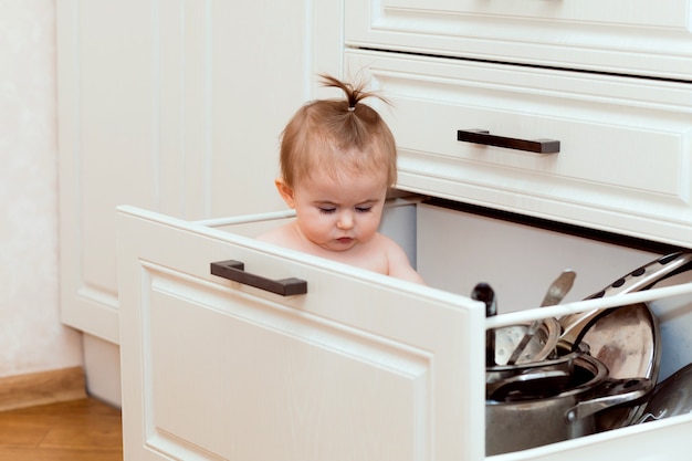 Niño feliz sentado en el cajón de la cocina con ollas y riendo. Retrato de un niño pequeño en una cocina blanca.