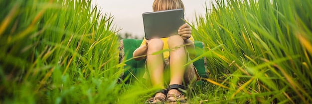 Niño feliz sentado en el campo sosteniendo tablet boy sentado en el césped en la escuela en casa día soleado