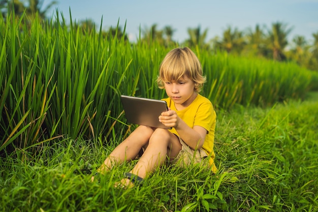 Niño feliz sentado en el campo sosteniendo una tableta Niño sentado en el césped en un día soleado Educación en el hogar o jugando una tableta