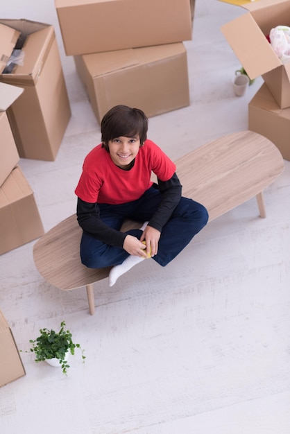 niño feliz sentado en la mesa con cajas de cartón a su alrededor en una nueva casa moderna, vista superior