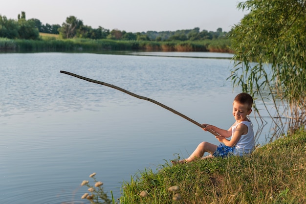 Un niño feliz sentado en la orilla de un estanque juega a pescar con un palo en sus manos