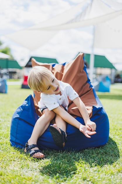 Niño feliz sentado en una silla de bolsa de frijoles en el parque de verano Un niño pequeño con una camiseta blanca se divierte al aire libre Infancia y felicidad