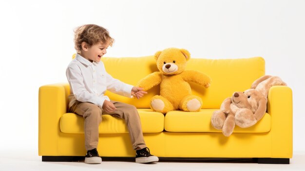 Niño feliz sentado en el sofá amarillo y dando un regalo al oso de peluche marrón aislado sobre un fondo blanco