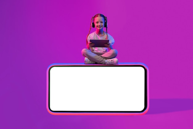 Niño feliz sentado en el teléfono con maqueta usando auriculares de tableta