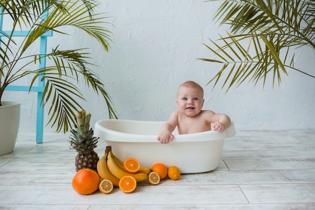 Niño feliz se sienta en una tina blanca con frutas tropicales sobre una superficie blanca con plantas con espacio para texto
