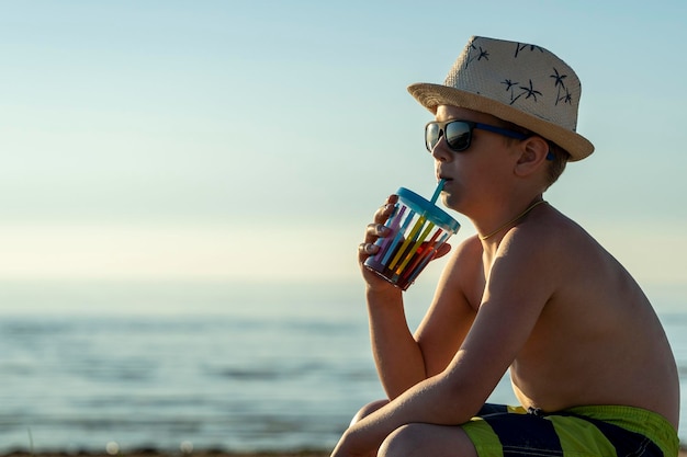 Niño feliz con sombrero y gafas de sol bebiendo jugo en la playa