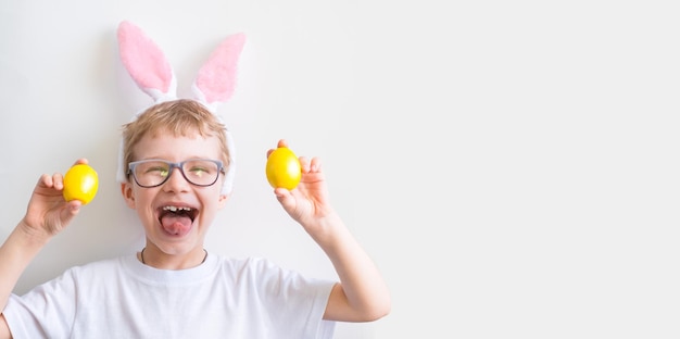 Niño feliz sonríe con gafas en una camiseta blanca en orejas de conejo y con coloridos huevos de Pascua amarillos