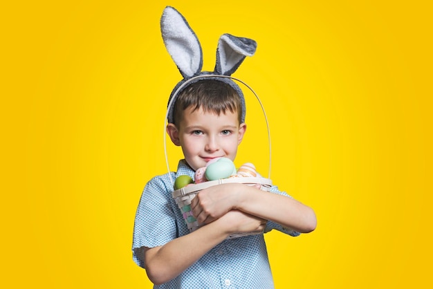 Un niño feliz y sonriente con orejas de conejo de Pascua sostiene una canasta con huevos pintados