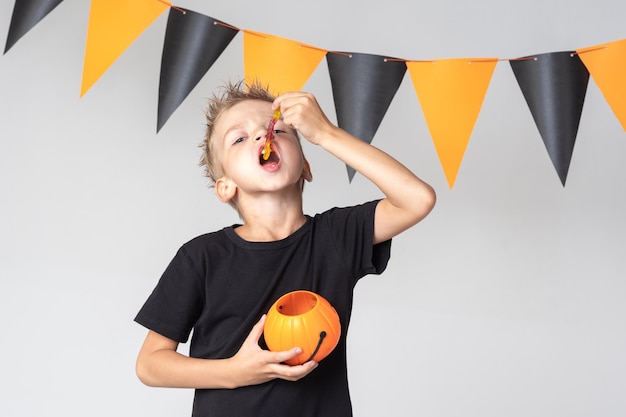 Un niño feliz y sonriente sostiene una canasta de Halloween con forma de calabaza y come mermelada de caramelo