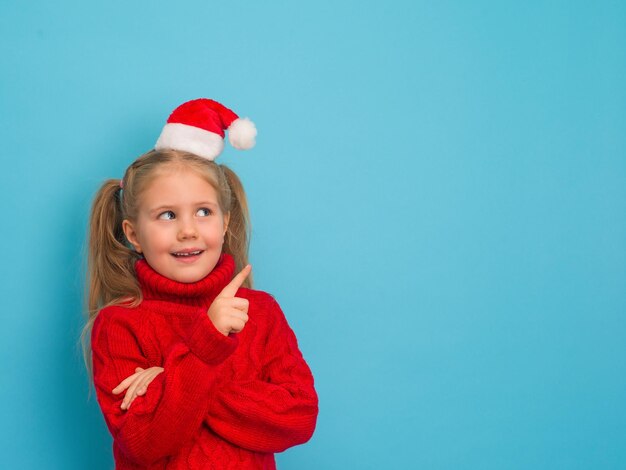 Niño feliz sorprendido con sombrero de Papá Noel y suéter rojo apuntando al espacio vacío de la copia en el fondo azul