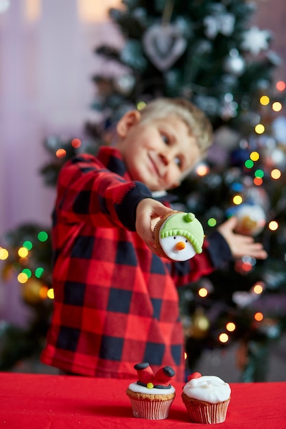 Niño feliz sosteniendo cupcakes creativos frente al árbol de Navidad Acogedor tiempo de Navidad Buen humor de vacaciones Enfoque selectivo