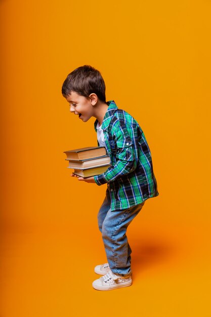 Niño feliz sosteniendo libros pesados aislado fondo amarillo. Concepto de educación