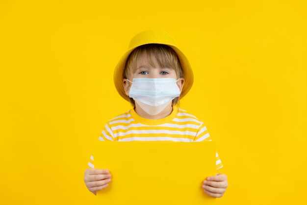 Niño feliz sosteniendo papel en blanco con máscara protectora interior. Vacaciones de verano durante la pandemia del coronavirus COVID-19.