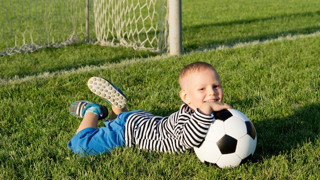 Foto niño feliz con su balón de fútbol tumbado sobre la exuberante hierba verde de un campo de deportes de verano en la luz del sol de la tarde