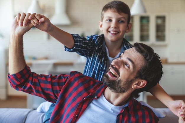 El niño feliz y su padre alegre se dan la mano de una manera juguetona y los dispersan a los lados sentados en el sofá de casa.