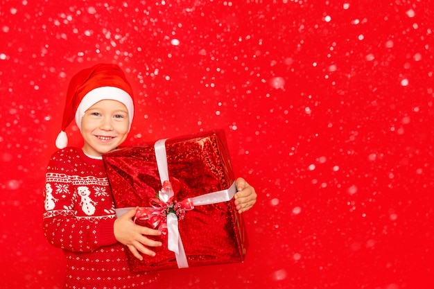 Un niño feliz con un suéter y gorro de Papá Noel se encuentra sobre un fondo rojo aislado con un gran regalo, espacio para texto