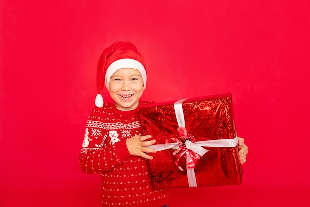 Un niño feliz con un suéter y gorro de Papá Noel se encuentra sobre un fondo rojo aislado con un gran regalo