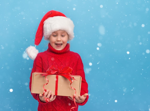 Niño feliz en suéter rojo y gorro de Papá Noel con regalo de Navidad sobre fondo azul.