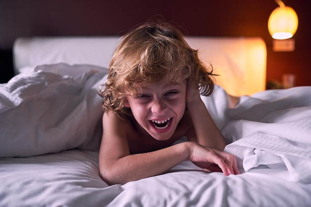 Un niño feliz tocando el cabello y riéndose de una broma mientras descansa bajo una manta en una cama suave en el hotel