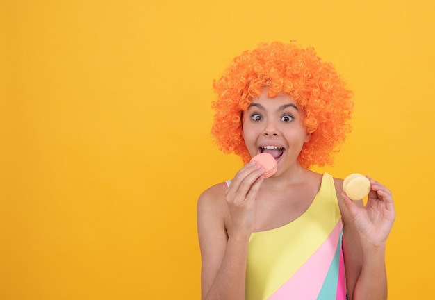 Niño feliz en traje de baño con pelo de peluca rizado naranja comiendo macaron francés sobre fondo amarillo, cuidado dental.