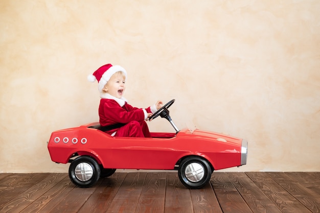 Niño feliz con traje de Santa Claus jugando en casa. Niño divertido conduciendo el coche de juguete. Concepto de vacaciones de Navidad