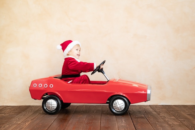 Niño feliz con traje de Santa Claus jugando en casa. Niño divertido conduciendo el coche de juguete. Concepto de vacaciones de Navidad