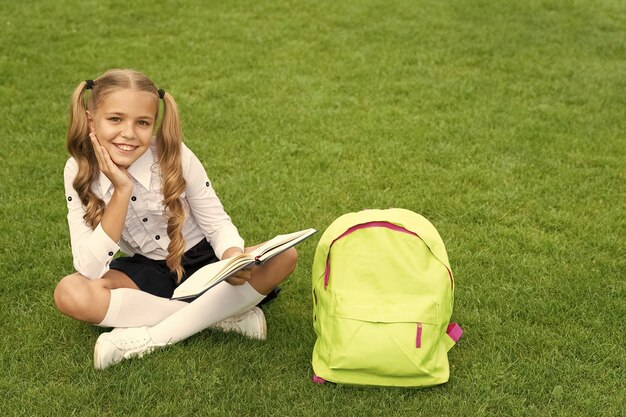 Niño feliz en uniforme con libro de lectura de mochila escolar sentado en la hierba verde