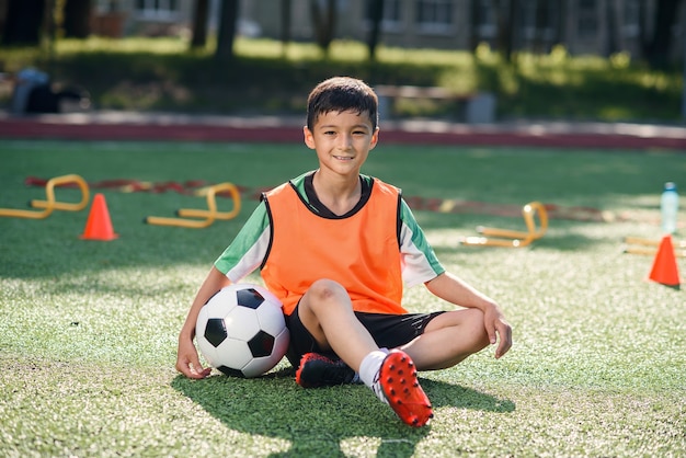 Niño feliz en uniforme sentado en el campo de fútbol con una pelota