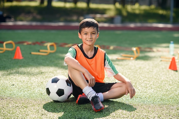 Niño feliz en uniforme sentado en el campo de fútbol con una pelota