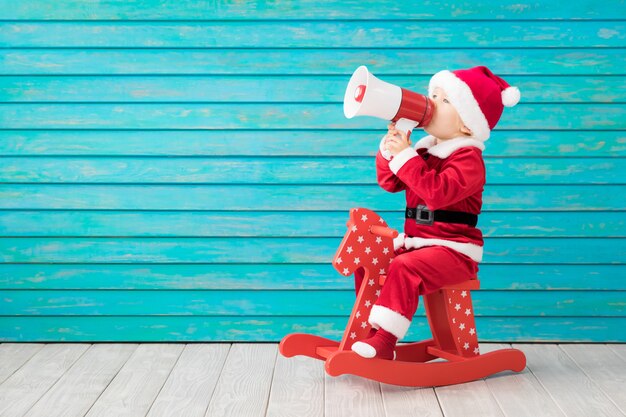 Niño feliz vestido con traje de Santa Claus jugando en casa. Niño divertido conduciendo un coche de juguete y hablando por megáfono. Concepto de vacaciones de Navidad