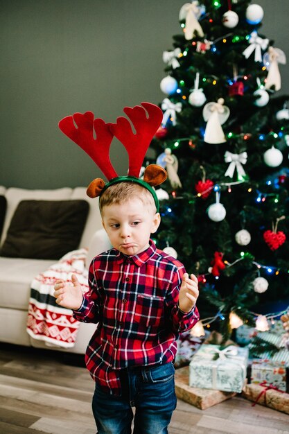 Niño feliz se viste en la cabeza de las astas de un ciervo cerca del árbol de Navidad.