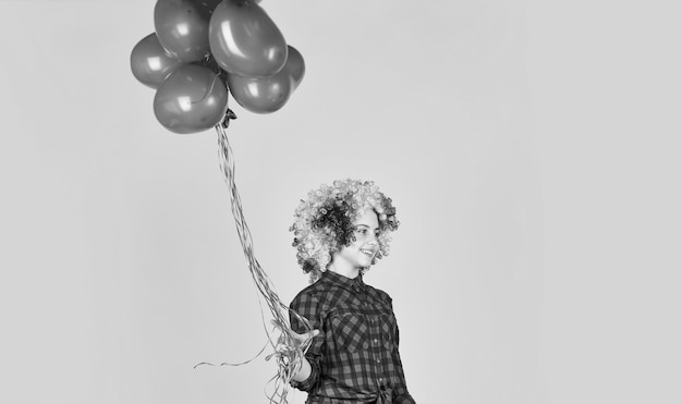 El niño de la fiesta del día de San Valentín usa la peluca del arco iris, el cabello, el fondo amarillo, el niño feliz, sostiene globos de fiesta, globos de corazón para el día del amor, sorpresa para San Valentín, niña pequeña colorida y brillante con peluca.