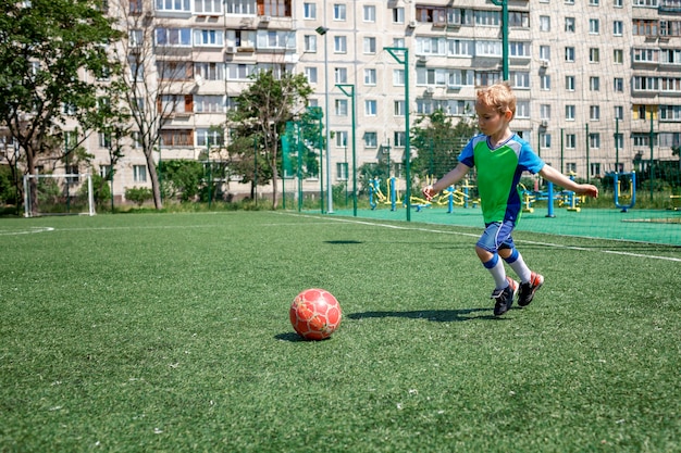 Niño en forma azul y verde jugando al fútbol en campo abierto en el patio de un joven jugador de fútbol