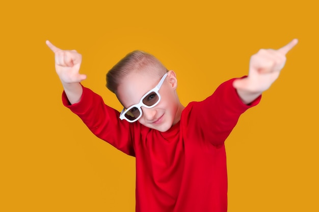 Un niño fresco y alegre con ropa roja y gafas muestra gestos con las manos sobre un fondo amarillo